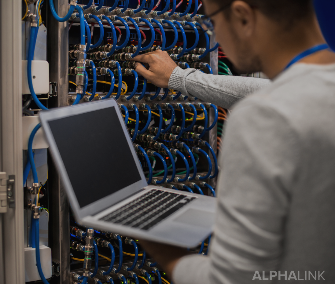 Técnico de ALPHALINK trabajando en un rack de red mientras revisa una laptop